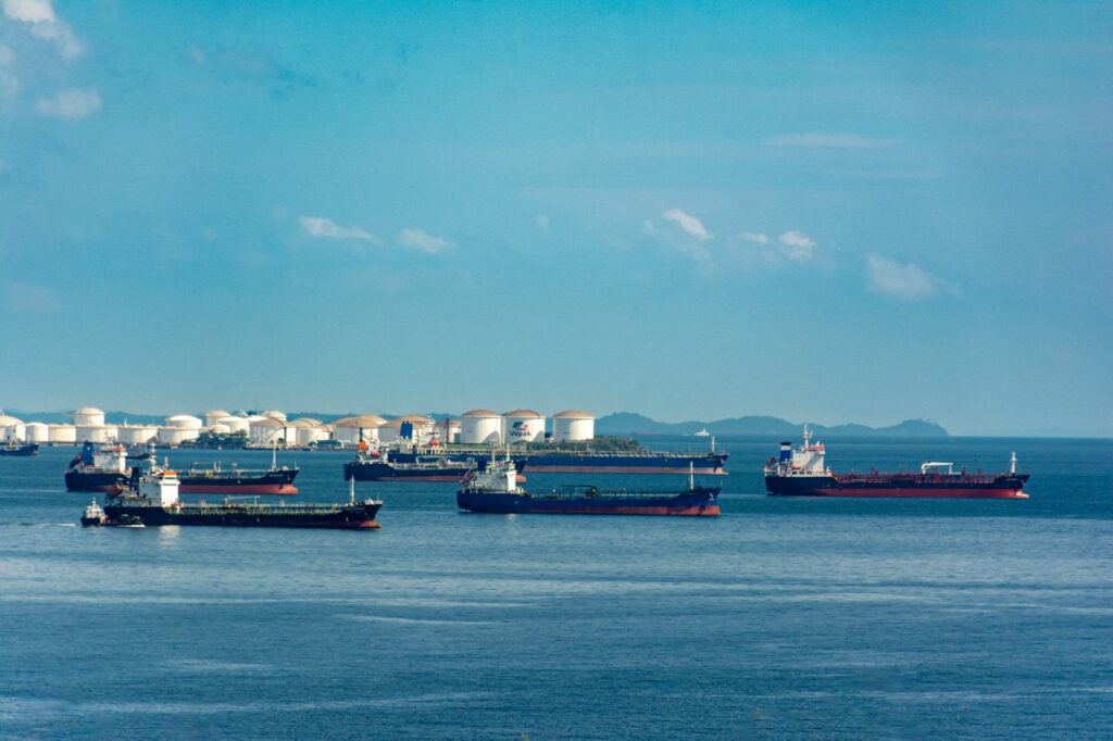 A fleet of cargo ships docked near oil storage tanks along a serene coastline with a clear blue sky above.