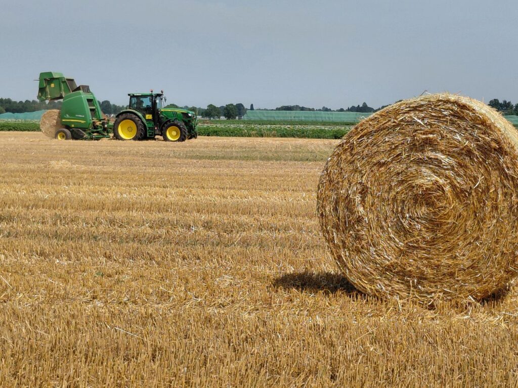 A tractor rolls hay bales in a vast field under a clear sky in Neuss, Germany, during summer.